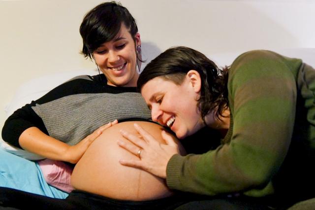A pregnant woman lays back, smiling, with her belly exposed, with her wife also smiling, placing her hand and head on the pregnant belly