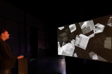 installation view of Neither Contradictory nor Coincidental with a person using a mouse on a pedestal in a dark room to control the work on a screen
