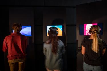 installation view of Videogames after Poetry with three people facing away from the camera each looking at an individual screen with headphones on