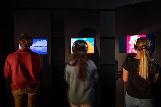 installation view of Videogames after Poetry with three people facing away from the camera each looking at an individual screen with headphones on