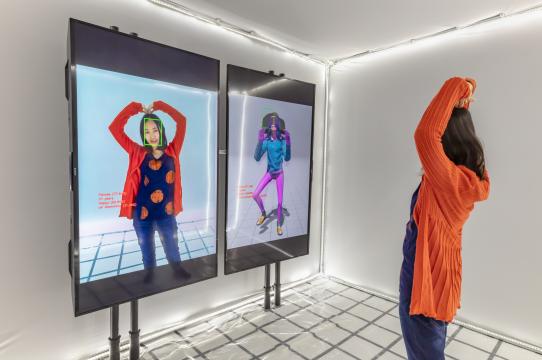 installation view of classification cube with a person holding their arms over their head in a white room lit from above and below with a gridded floor and two screens in front of them