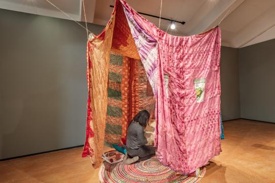 installation view of stitching solidarity with person kneeling inside hung tent of quilts