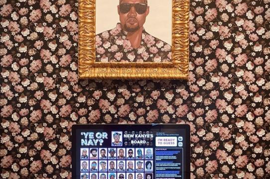Desktop computer in a gallery installation, showing digital game Ye or Nay, in front of a floral wallpaper and portrait of Kanye west in matching floral shirt