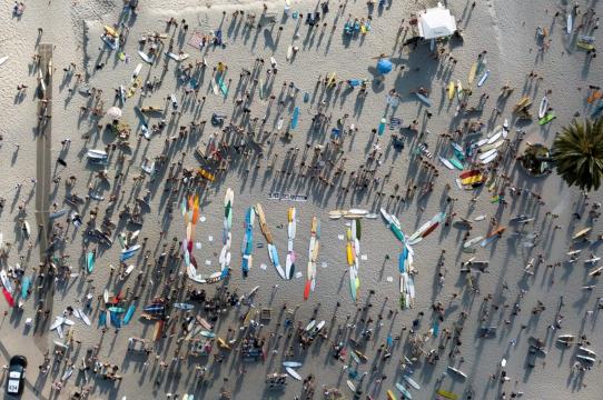 Overhead photos of surfboards laid out on the beach spelling the word UNITY surrounded by a crowd of people